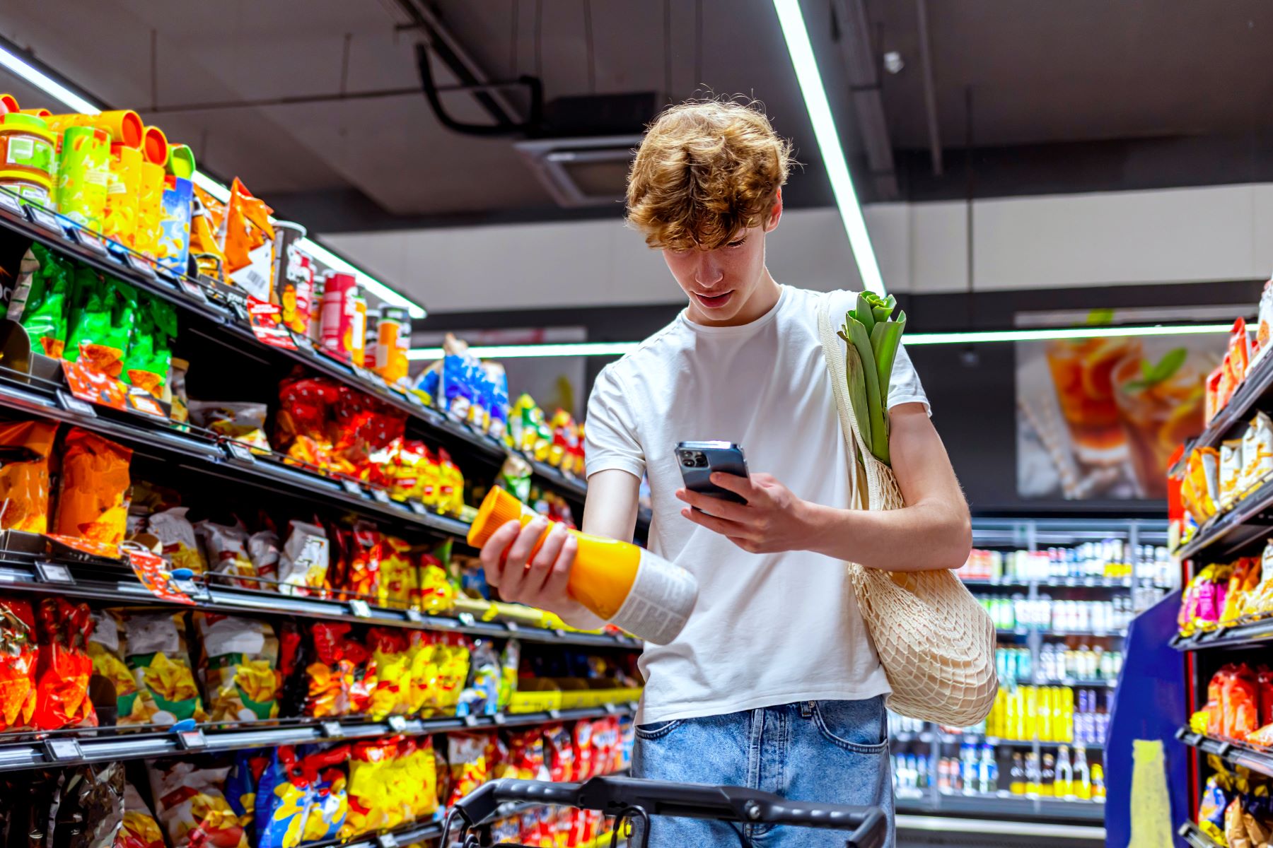 Young man in white t-shirt holding a bottle and using a smartphone while grocery shopping in a store aisle filled with colorful packaged snacks.